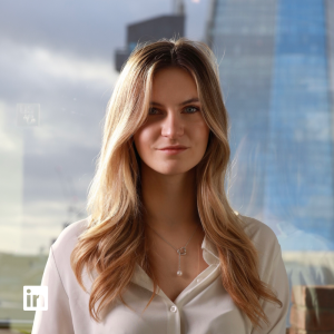 Portrait of a woman with long blonde hair wearing a white blouse, standing in front of a glass skyscraper and posing for a professional photo.