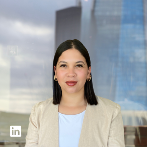 Professional woman portrait for LinkedIn: woman with straight dark hair, beige blazer, light blue blouse, looking at the camera against a glass-building background.