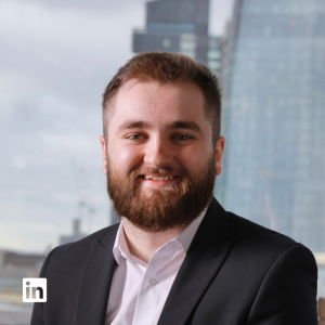 Smiling bearded man in a black suit and white shirt with an urban skyline behind him; LinkedIn badge in the lower-left corner.