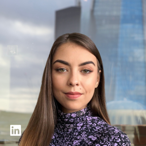Professional headshot of a young woman with long brown hair, wearing a purple floral turtleneck, standing in front of a glass building (LinkedIn watermark).
