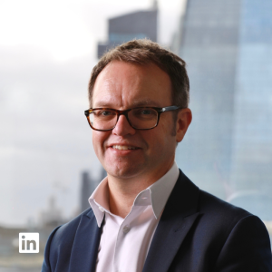 Smiling man in glasses wearing a navy blazer and white shirt, posing for a professional headshot with a cityscape background and a LinkedIn logo overlay.
